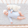 A baby in a blue onesie lies in a Makemake Organics crib with a Cobi Blue Stripes fitted sheet and pillowcase, accompanied by a plush teddy bear on its side. The baby is looking up and smiling, wearing a white headband.
