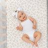 A happy baby wearing a white onesie and matching bow headband lies on a Makemake Organics Wild crib fitted sheet with pillowcase, featuring a beige animal print design, looking up and smiling.
