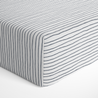 A corner view of a mattress covered with a Makemake Organics Cobi Blue Stripes crib fitted sheet. The sheet is neatly tucked, emphasizing the crisp, clean lines of the design.