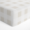 A close-up of a Makemake Organics crib fitted sheet with pillowcase in a checkerboard pattern in shades of light gray and cream. The corner is shown, highlighting the squared and slightly rounded shape of the mattress edge.