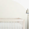 A minimalist nursery with a white Makemake Organics crib against a cream wall with a large beige half-circle painted on it. there's a floor lamp with a metallic finish to the right.