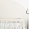 A minimalist nursery room featuring a white Makemake Organics baby crib with a Crib Fitted Sheet with Pillowcase - Safari bedding set, against a cream wall with a large half-circle paint design. A floor lamp is to the right.