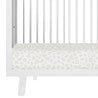 Close-up of a Makemake Organics white crib with a mattress covered in a Wild leopard print design. The crib features vertical bars and a visible side rail.