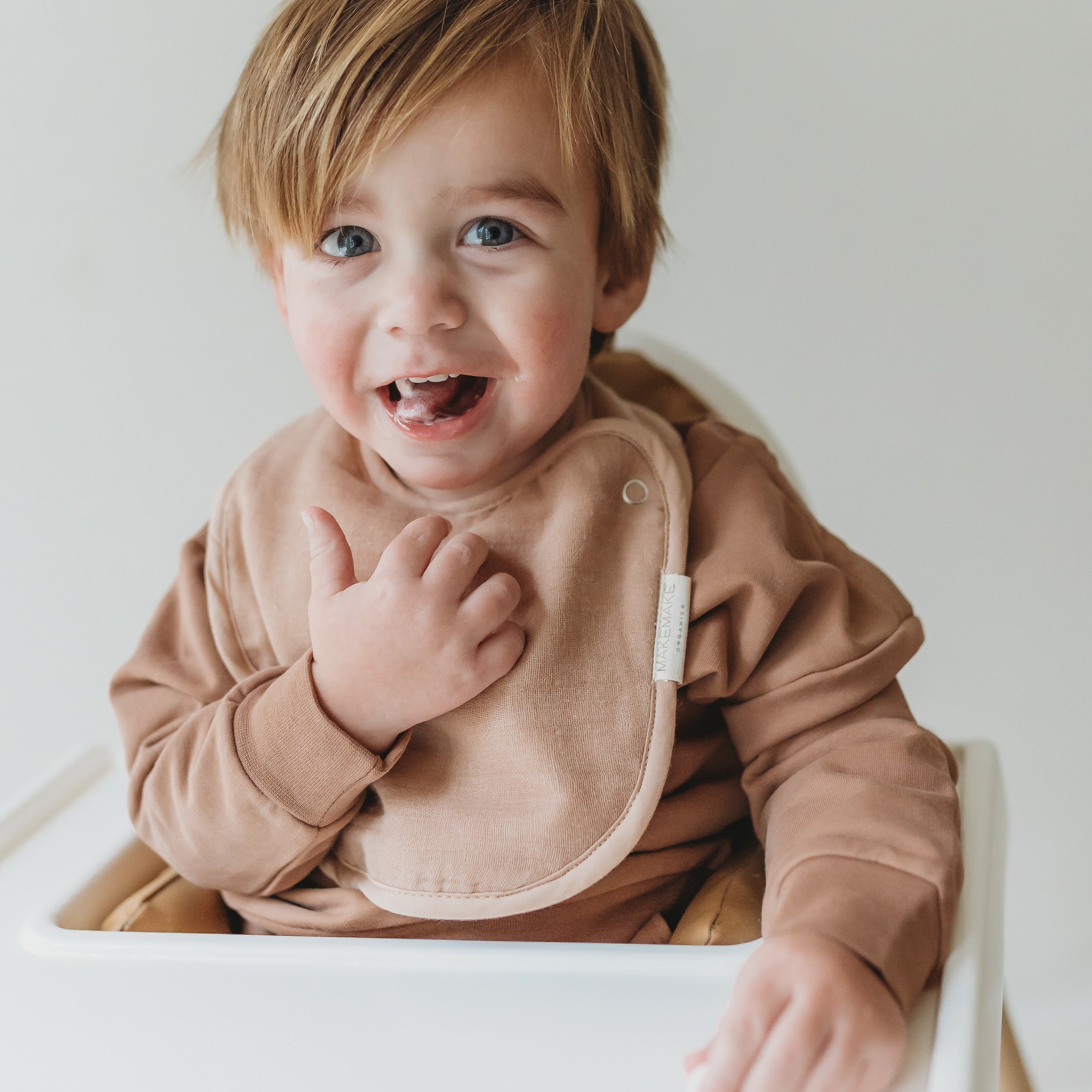 A cheerful toddler with tousled hair sitting in a white high chair, dressed in a brown long-sleeve shirt and Makemake Organics Organic Muslin Bibs - Blush Oat & Pecan, pointing at their chest with a joyful expression.