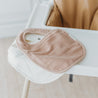 A light brown Makemake Organics Organic Muslin Bib rests on a white high chair in a brightly lit room, highlighting a simple and clean design.