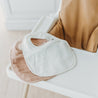 Three Organic Muslin Bibs in Ivory & Taupe from Makemake Organics displayed on a white high chair against a bright background, illustrating a clean, minimalist baby feeding setup.