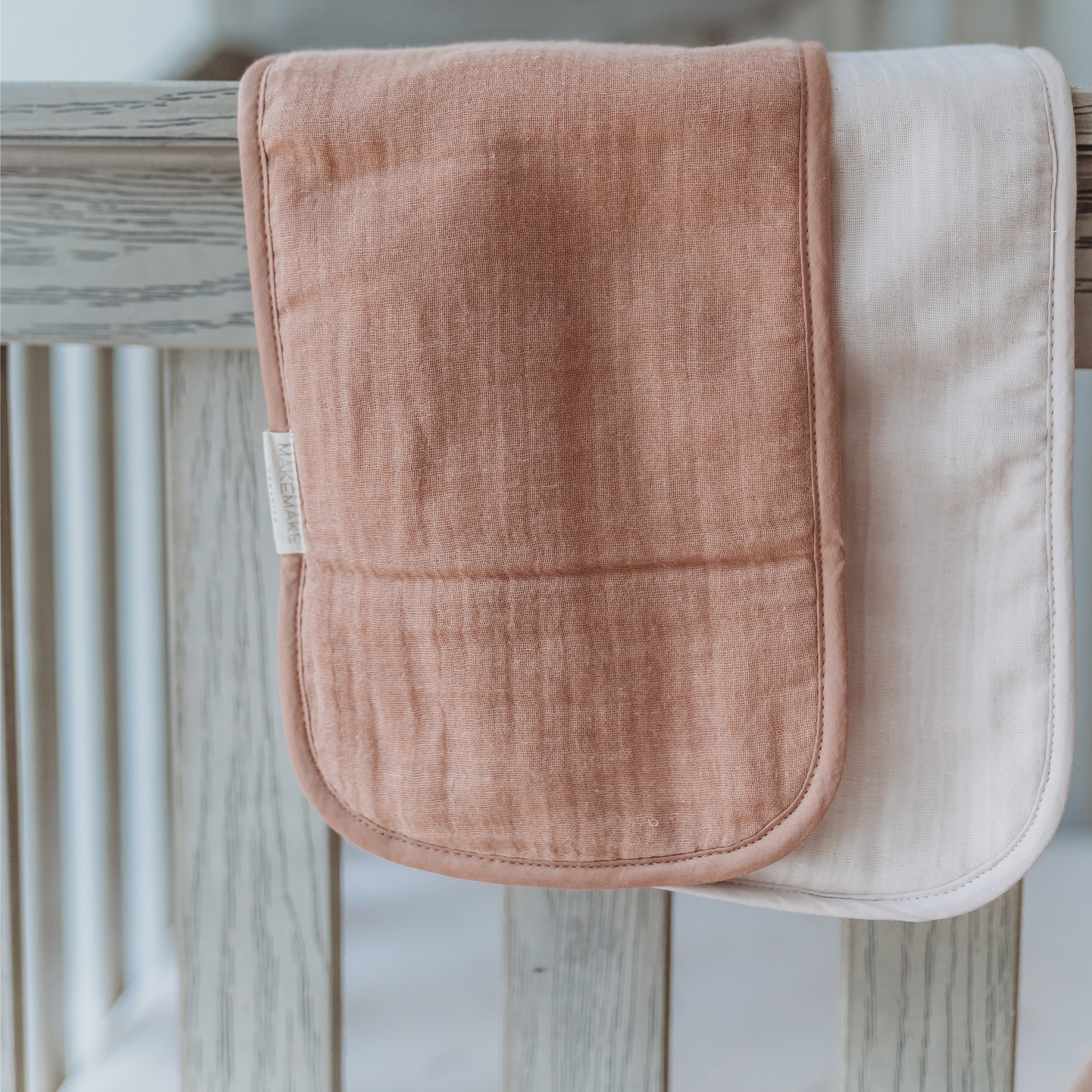 Two Organic Cotton Muslin Burp Cloths in Blush Oat & Pecan by Makemake Organics hanging on a wooden rail, one in a blush pink and the other in a soft white, both with textured surfaces.