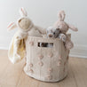 A Makemake Organics Storage Basket Pompom Blush Oat adorned with pink pom-poms, containing a plush bunny, a kitten, and a bunny-themed blanket, on a wooden floor against a white wall.