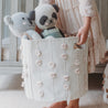 A child holds a Makemake Organics Storage Basket Pompom Blush Oat containing two plush toys, a bear and a panda, in a softly lit room.
