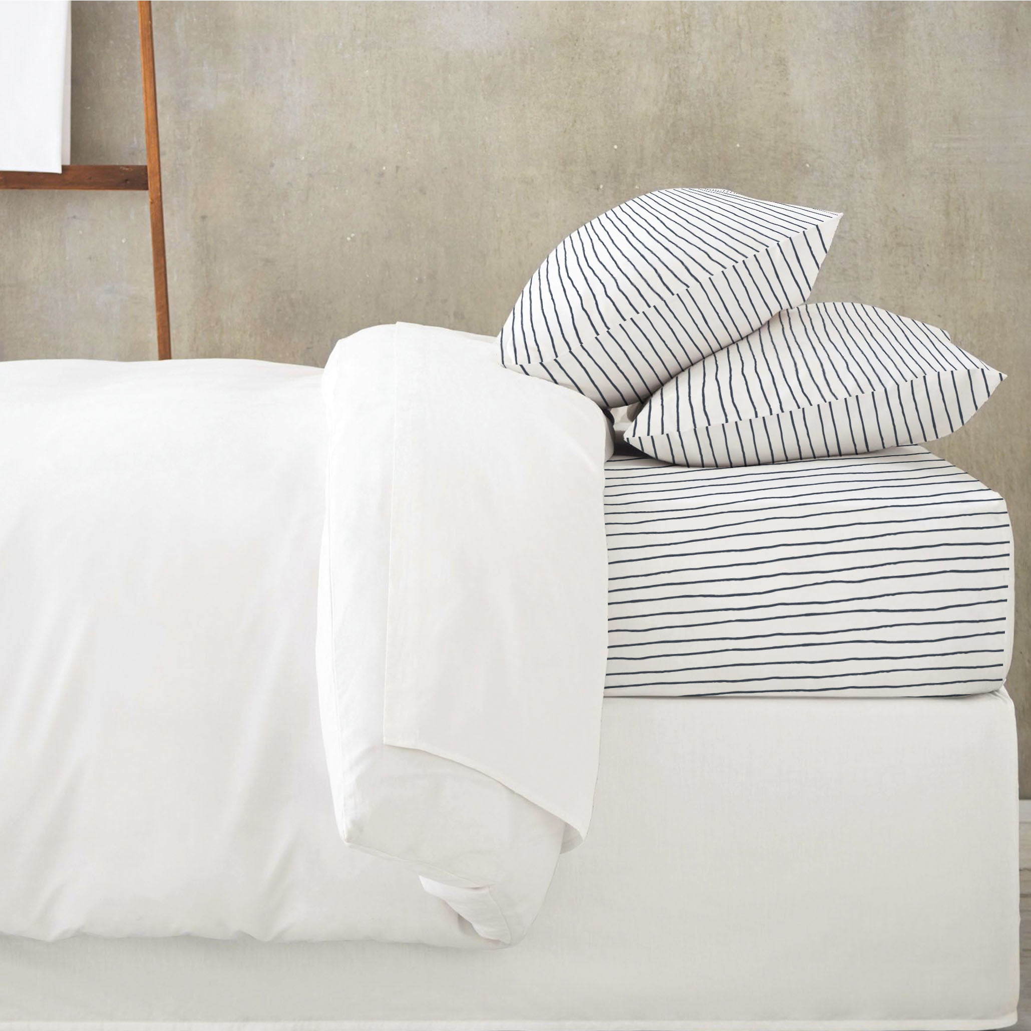 A neatly made bed with white bedding and Makemake Organics Organic Cotton Fitted Sheet Set in Navy Stripes against a muted gray background.