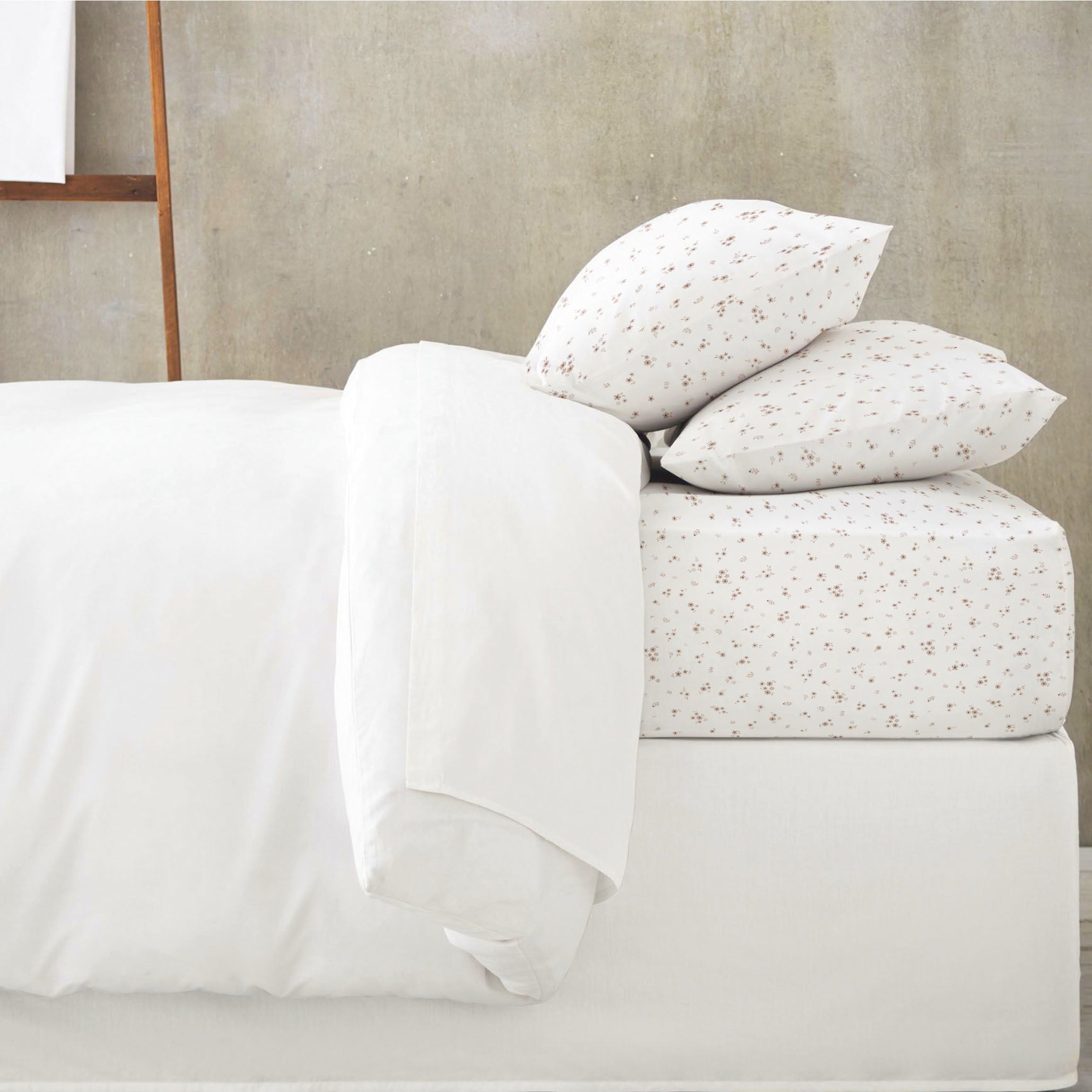 A neatly made bed with a white duvet and two pillows, one with a white cover and another with a tiny floral print, set against a grey wall.