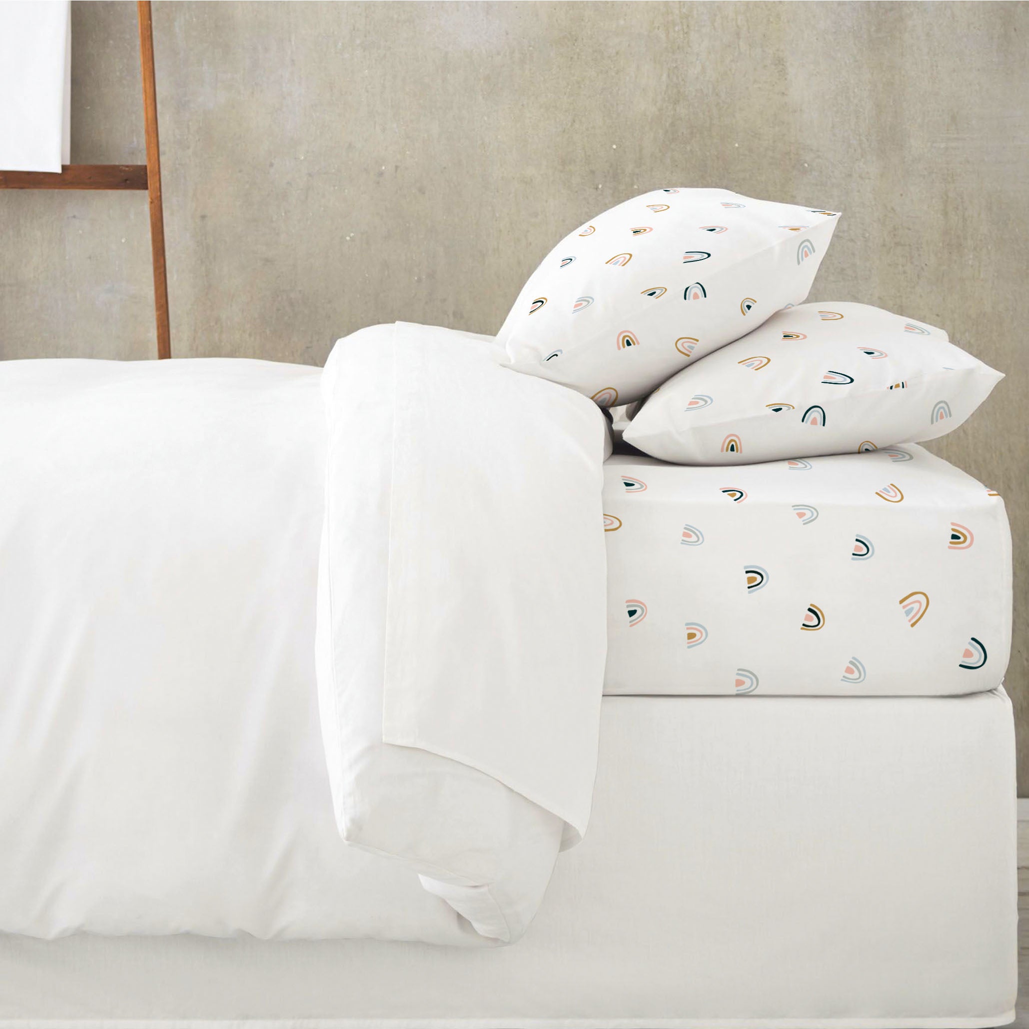 A neatly made bed with Makemake Organics' Organic Cotton Fitted Sheet Set - Rainbow, featuring a rainbows and moon design, positioned against a plain textured wall.