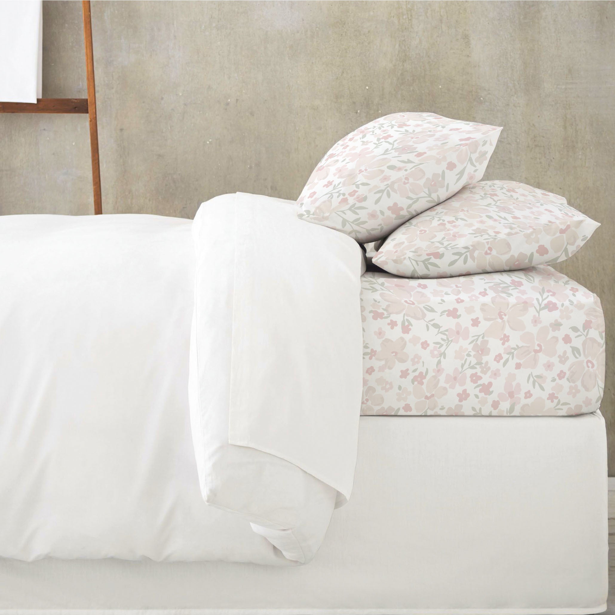 A neatly made bed with an Organic Cotton Fitted Sheet Set - Blossom from Makemake Organics against a gray concrete wall. Two pillows have a subtle floral pattern and the duvet is plain white.