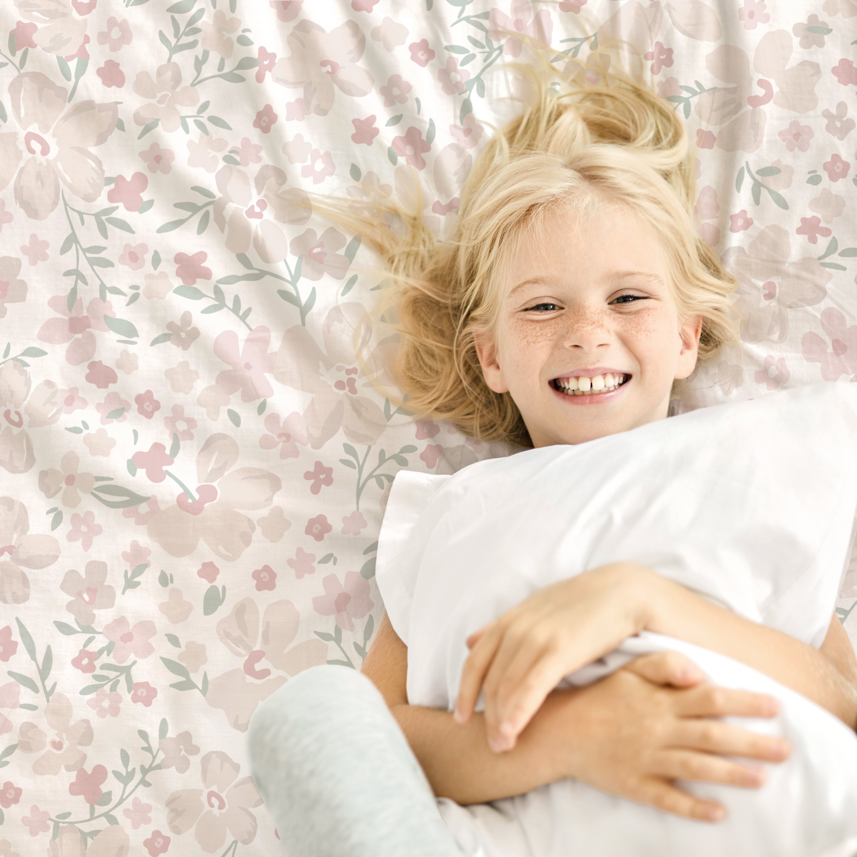 A young girl with blond hair smiling broadly, lying on an Organic Cotton Sheet Set - Blossom from Makemake Organics, hugging a white pillow, viewed from above.