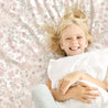 A joyful young girl with blonde hair smiling, lying on a Makemake Organics Organic Cotton Fitted Sheet Set - Blossom, and hugging a white pillow.