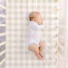 A newborn baby sleeps peacefully in a crib, dressed in a white onesie, lying on a back in a soft, striped Makemake Organics fitted sheet with pillowcase. The view is from above.