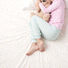 A young girl in pajamas, holding a teddy bear, sitting on a Makemake Organics Organic Cotton Fitted Sheet Set - Dotty with her legs crossed, seen from above.