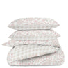 Five stacked pillows and a Makemake Organics Organic Duvet Cover - Blossom & Plaid, with floral and check patterns in soft pink and gray colors, isolated on a white background.