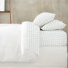 A neatly made bed with white and light gray checkered bedding, including a Makemake Organics Organic Cotton Sheet Set - Gingham, duvet, and four pillows against a concrete wall background.