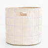 A blush oat fabric storage basket with visible zigzag stitching patterns, equipped with a small wooden label holder on the front, against a white background by Makemake Organics.