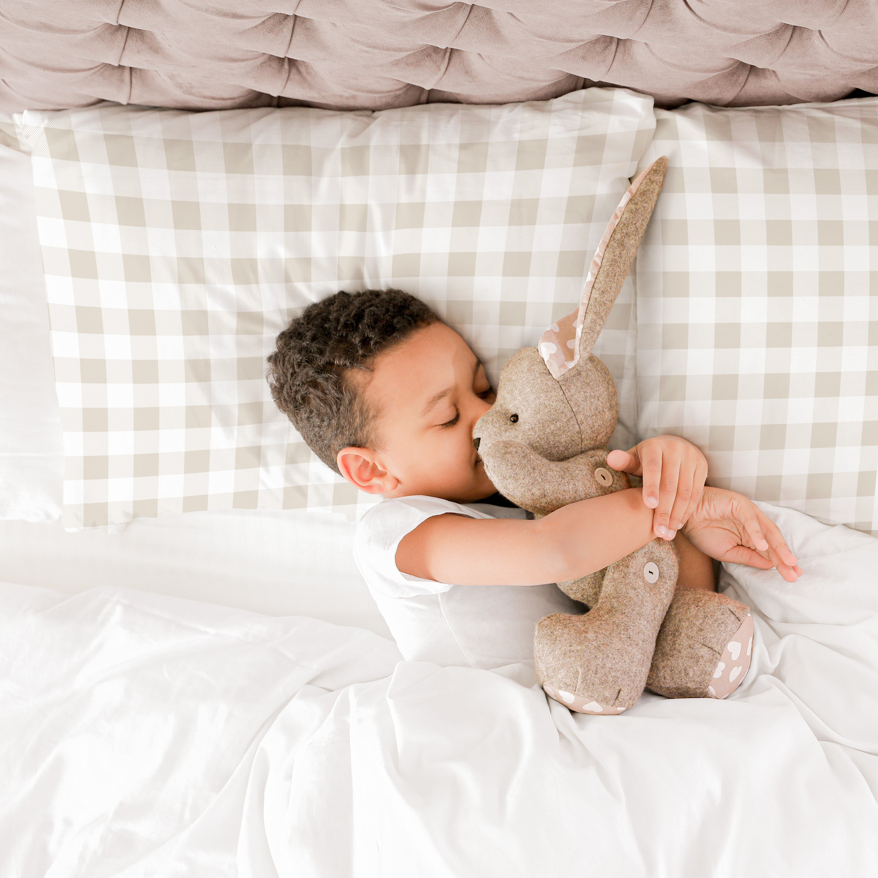 A young child lying on a bed, affectionately kissing a plush bunny toy. the bedding has a checkered pattern, creating a cozy atmosphere with the Makemake Organics Organic Cotton Toddler Pillowcase in Plaid.