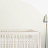 A minimalist nursery room with a white crib and a standing lamp in the corner, featuring a large pastel green half-circle painted on a light beige wall. The crib is adorned with Makemake Organics' Crib Fitted Sheet with Pillowcase - Plaid.