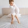 A joyful young boy in white pajamas playfully lifting a pillow while standing on a Makemake Organics Organic Cotton Fitted Sheet Set - Navy Stripes in a bright bedroom.
