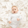 A smiling baby in a white onesie lies on a bed with sheets from Makemake Organics featuring a safari animal print, including zebras and birds.