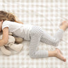 A young girl lying face down on a Makemake Organics plaid fitted sheet set, wearing a white shirt and striped leggings, clutching a brown teddy bear. Her blonde hair is spread out.