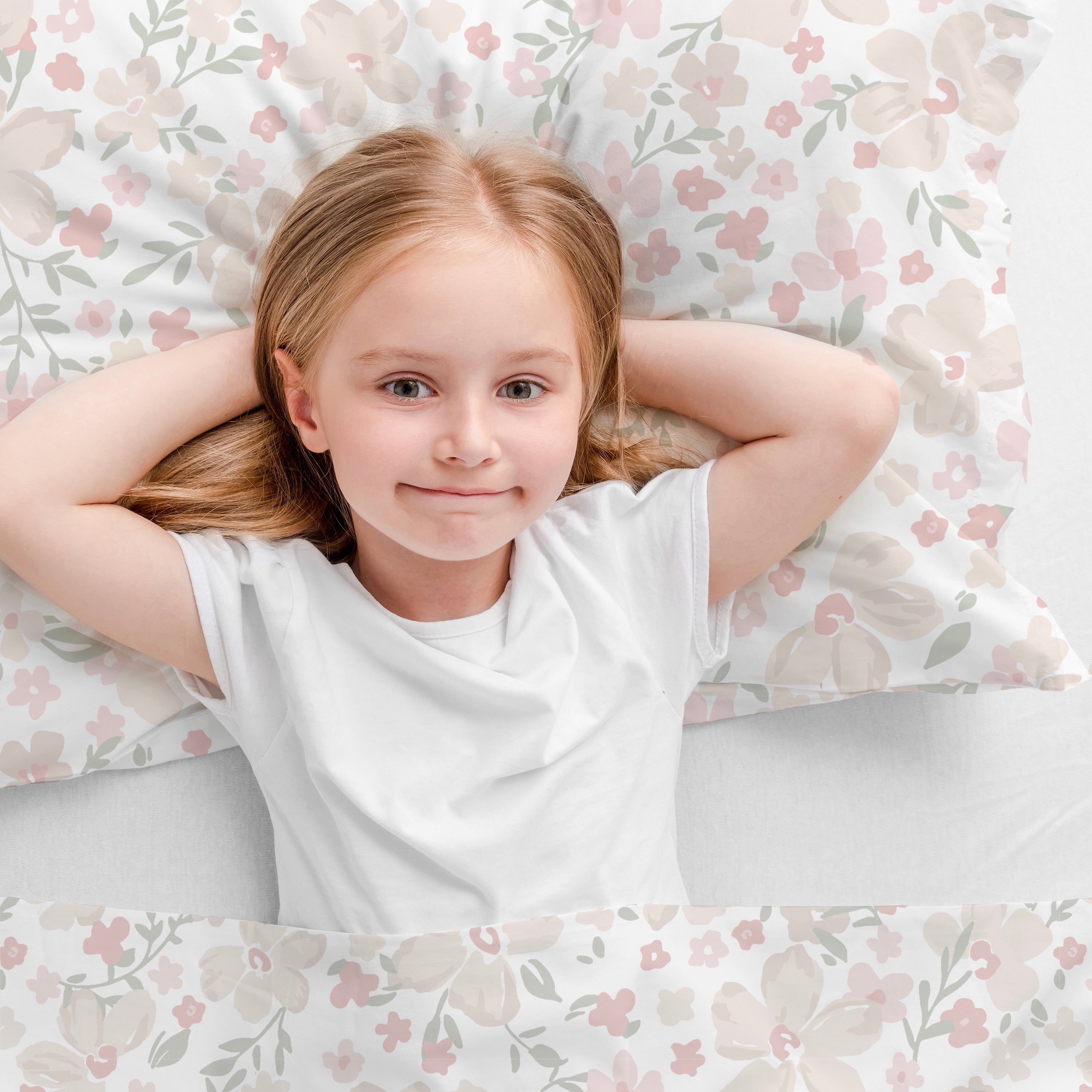 A young girl with light hair and a gentle smile, lying on her back on a bed with Makemake Organics Organic Cotton Toddler Pillowcase in Blossom print bedding, hands resting behind her head, looking up at the camera.