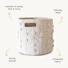A collapsible, sturdy canvas storage bin with handles, featuring a beige color and adorned with pink pompoms. it includes labels highlighting its easy handling, ethical production, and collapsible fabric design. The Storage Basket Pompom Blush Oat by Makemake Organics is a stylish and functional addition to any space.