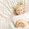 A joyful young girl with blonde hair, lying on a bed with a Makemake Organics Organic Cotton Fitted Sheet Set - Wild duvet, smiling at the camera while clutching a white pillow.