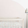 A minimalist nursery with a white crib from Makemake Organics featuring their Blossom fitted sheet and pillowcase, positioned against a soft pink half-circle painted wall. A floor lamp stands to the side.