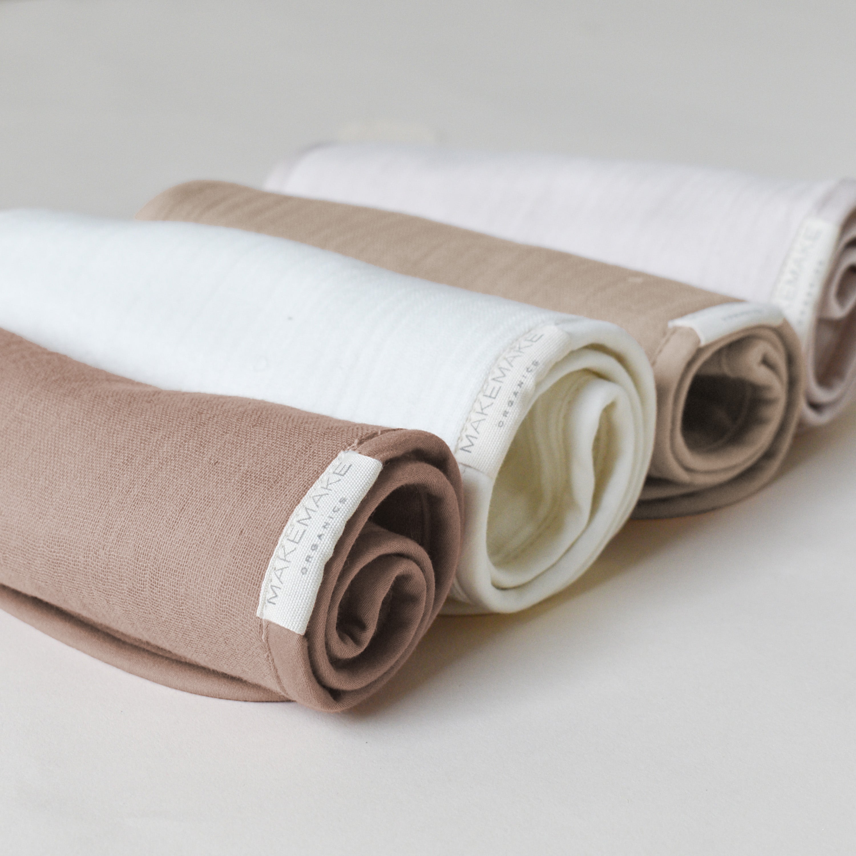 Three rolled fabrics in white, tan, and brown on a neutral background, each labeled with the brand "Makemake Organics Organic Cotton Muslin Burp Cloth - Ivory & Taupe.