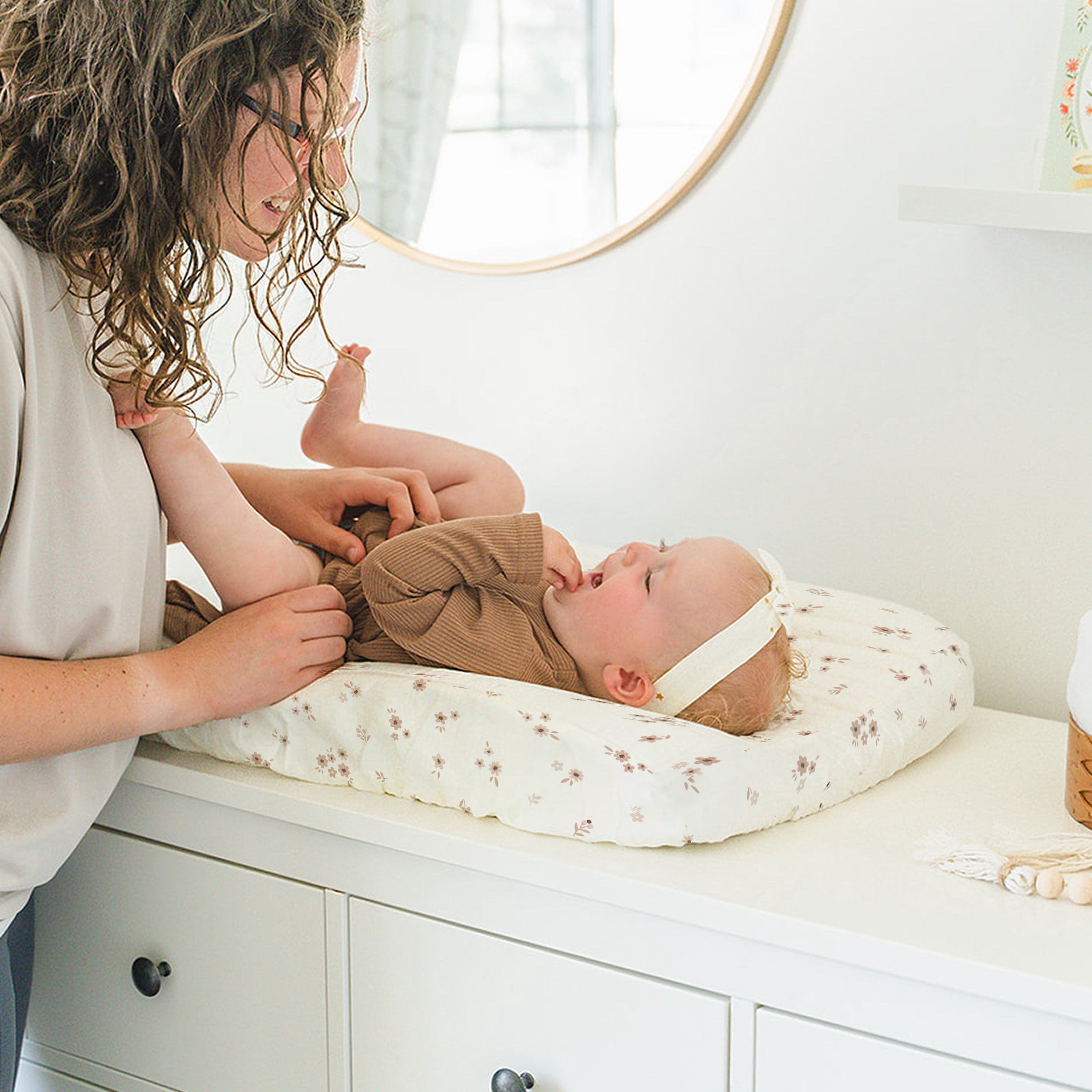 A young girl gently touches a baby who is lying on a Organic Cotton Changing Pad Cover by Makemake Organics on a white dresser in a bright, neutrally decorated room.