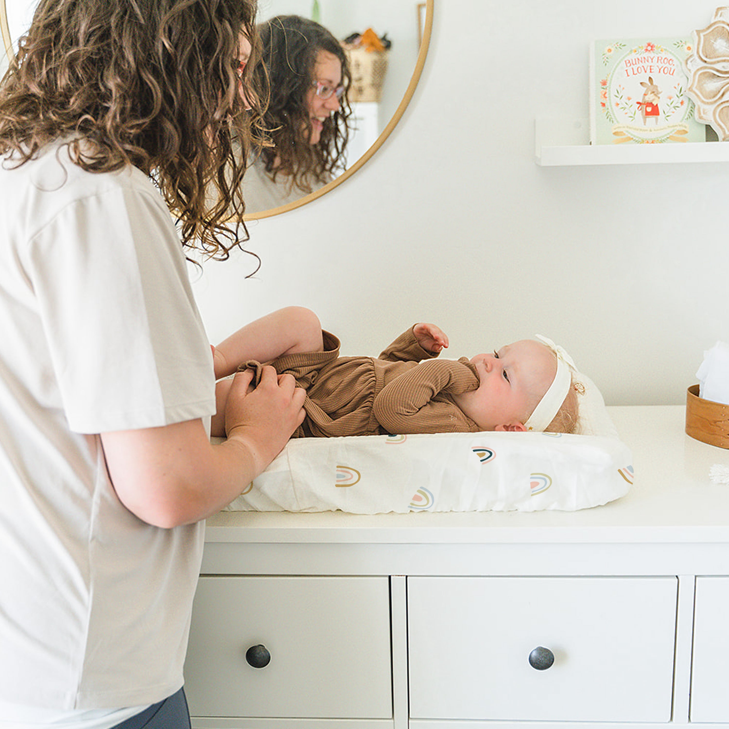 A woman with curly hair, in a white t-shirt, gently interacts with a baby lying on a changing table in a bright, well-organized nursery featuring the Makemake Organics Organic Cotton Changing Pad Cover - Rainbow.