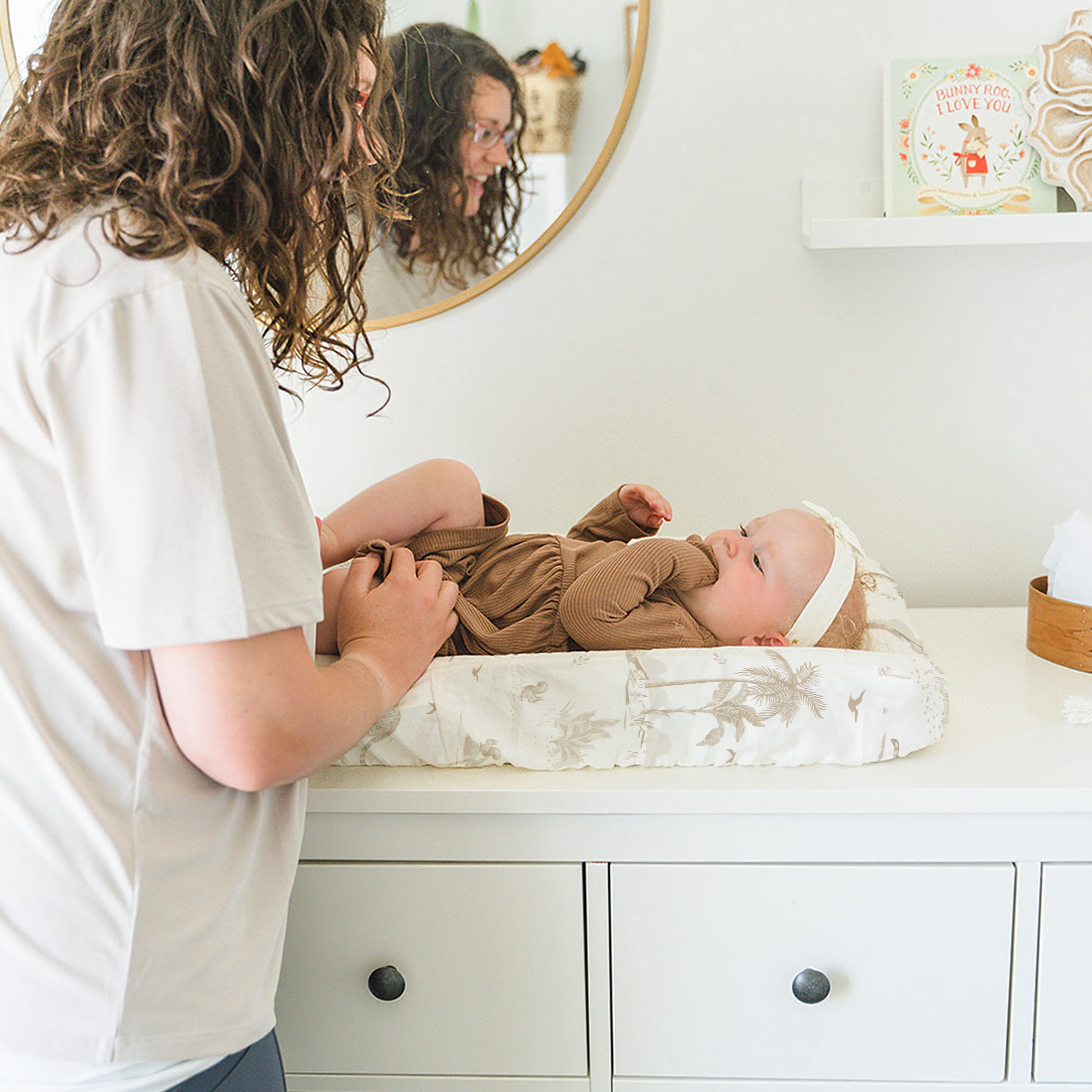 A woman engaging with a baby lying on a Makemake Organics Organic Cotton Changing Pad Cover - Safari atop a white dresser. The room is bright, and there's a mirror reflecting the duo's interaction.