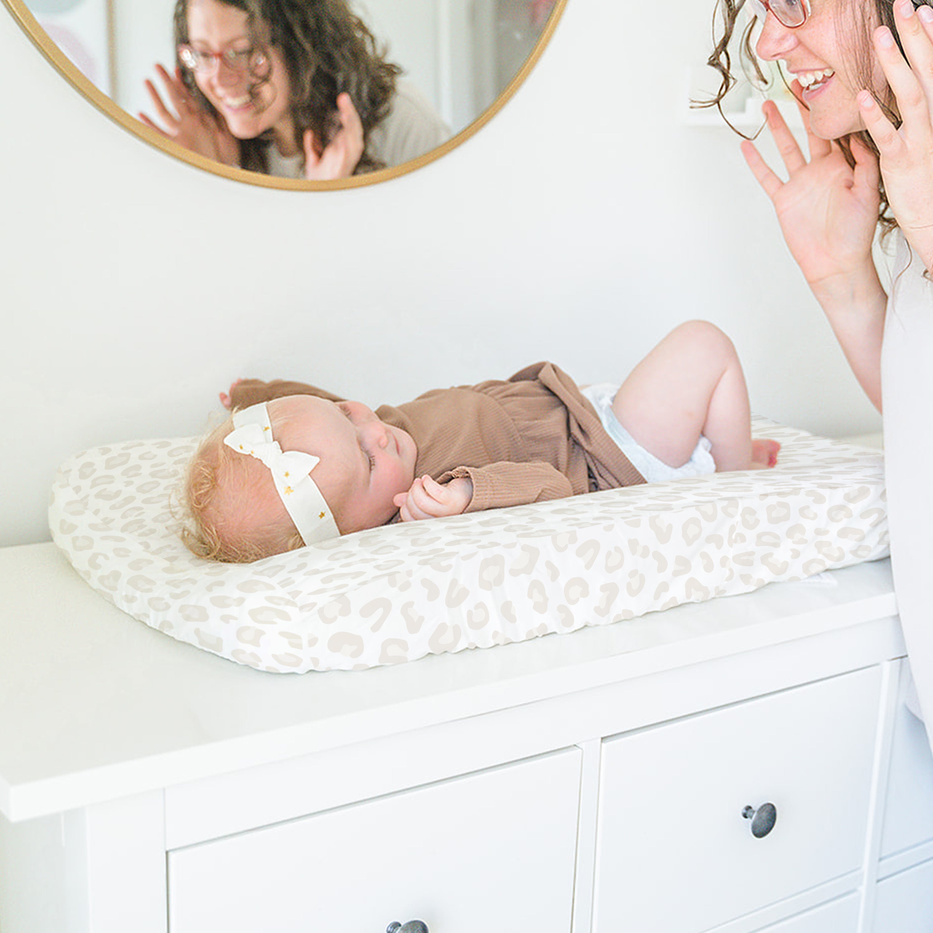 A mother gazes happily at her baby, who is lying on a Makemake Organics Organic Cotton Changing Pad Cover - Wild, under a round mirror reflecting both their cheerful expressions.