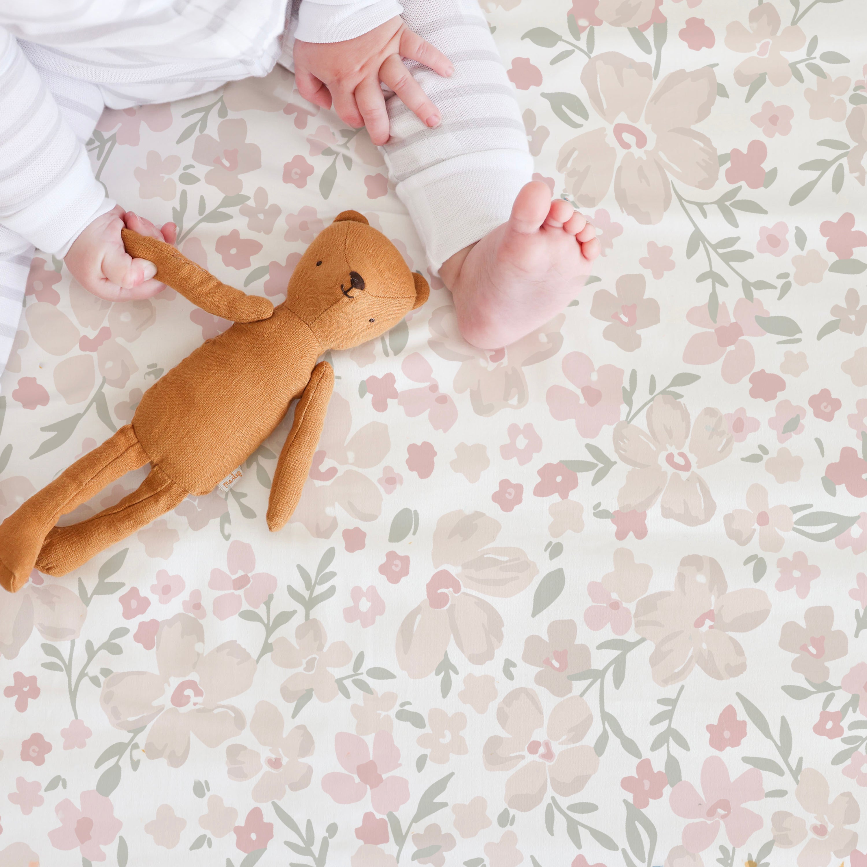 A baby's legs and arms are visible as they hold a Makemake Organics Blossom crib fitted sheet with pillowcase on a floral patterned blanket with soft pink and green colors.