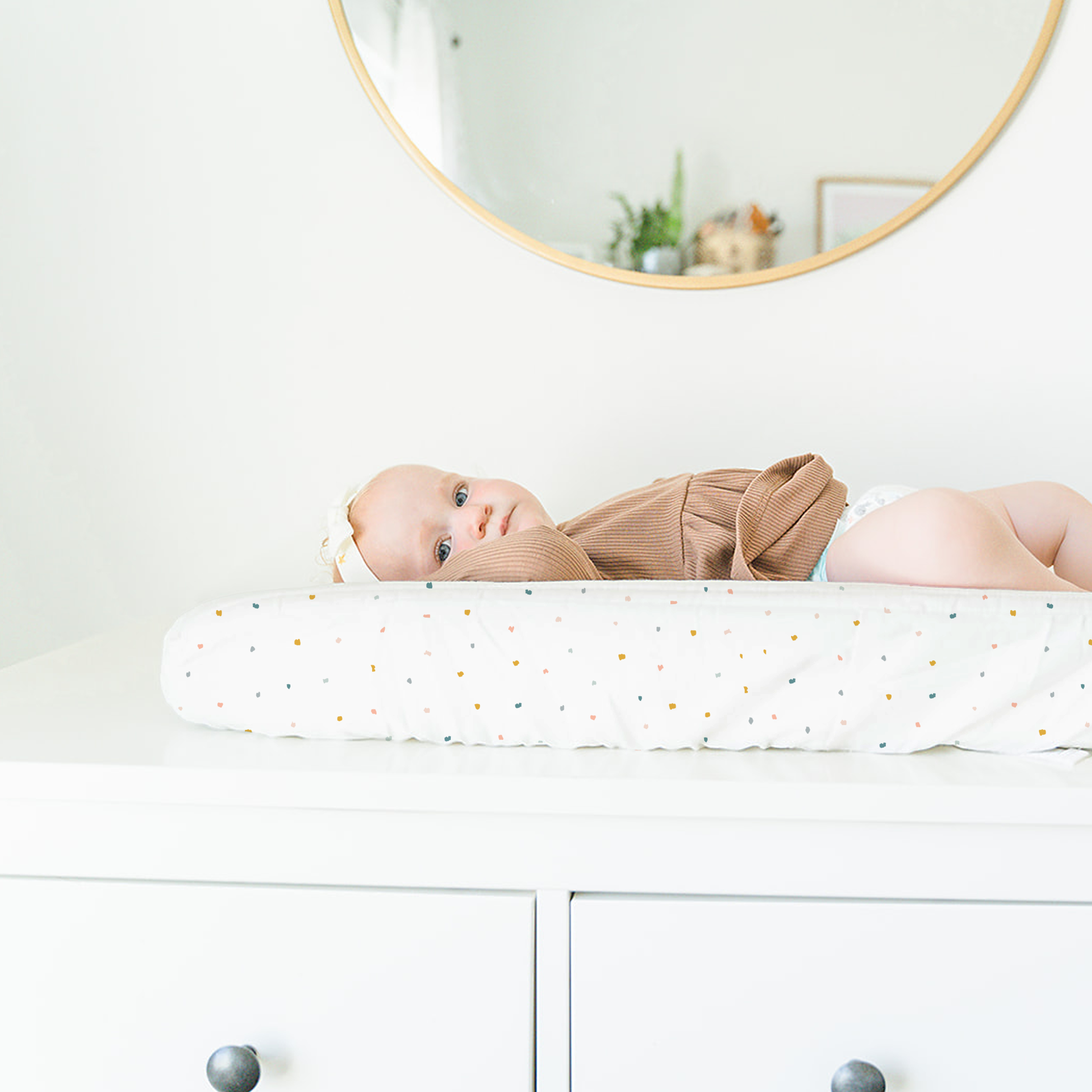 A baby wearing a brown outfit lying on a Makemake Organics Organic Cotton Changing Pad Cover - Dotty, looking up, with a round mirror overhead reflecting a light interior.