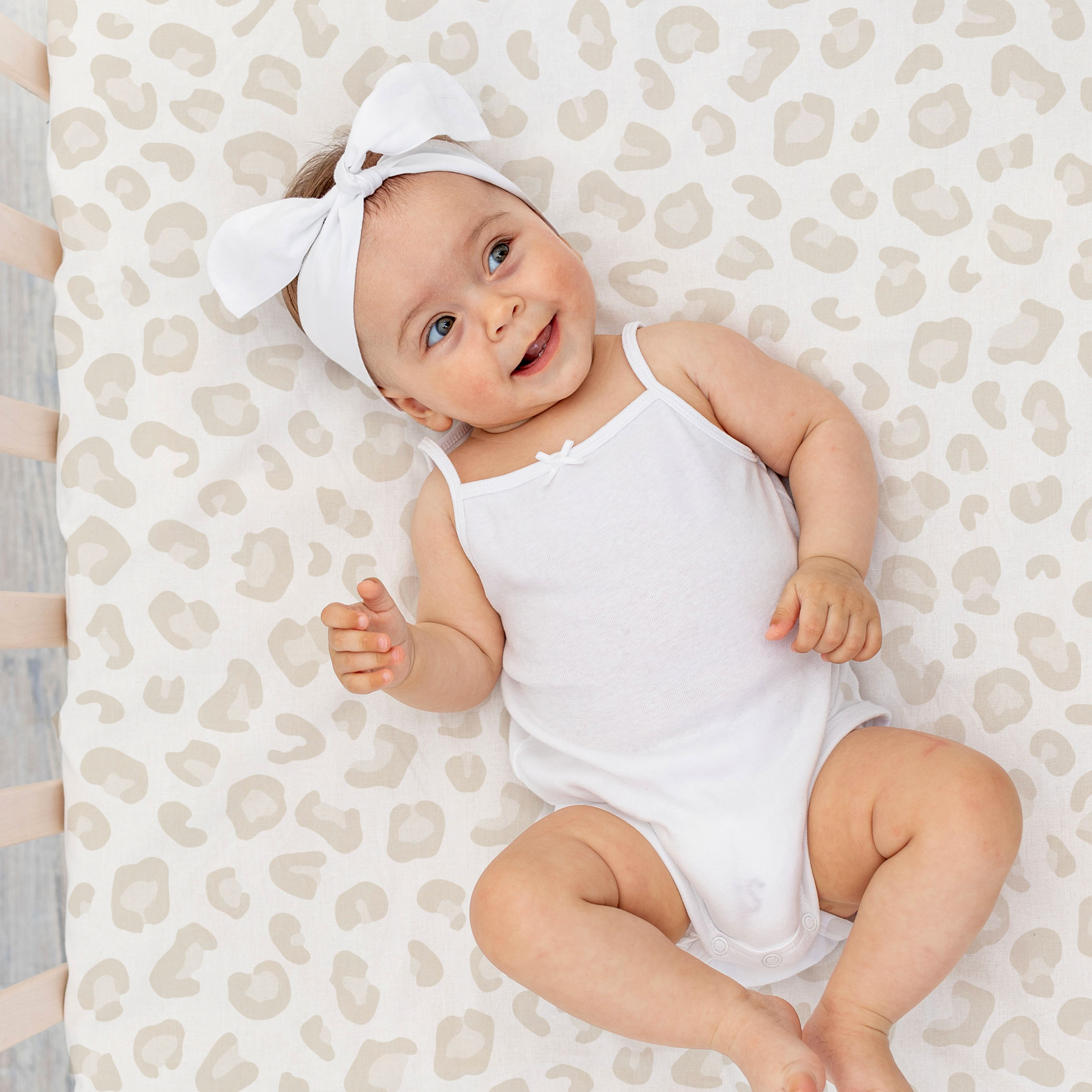 A smiling baby wearing a white onesie and a matching headband lies on a patterned blanket, looking upward with joy on a Mini Crib Fitted Sheet Set- Safari & Wild from Makemake Organics.