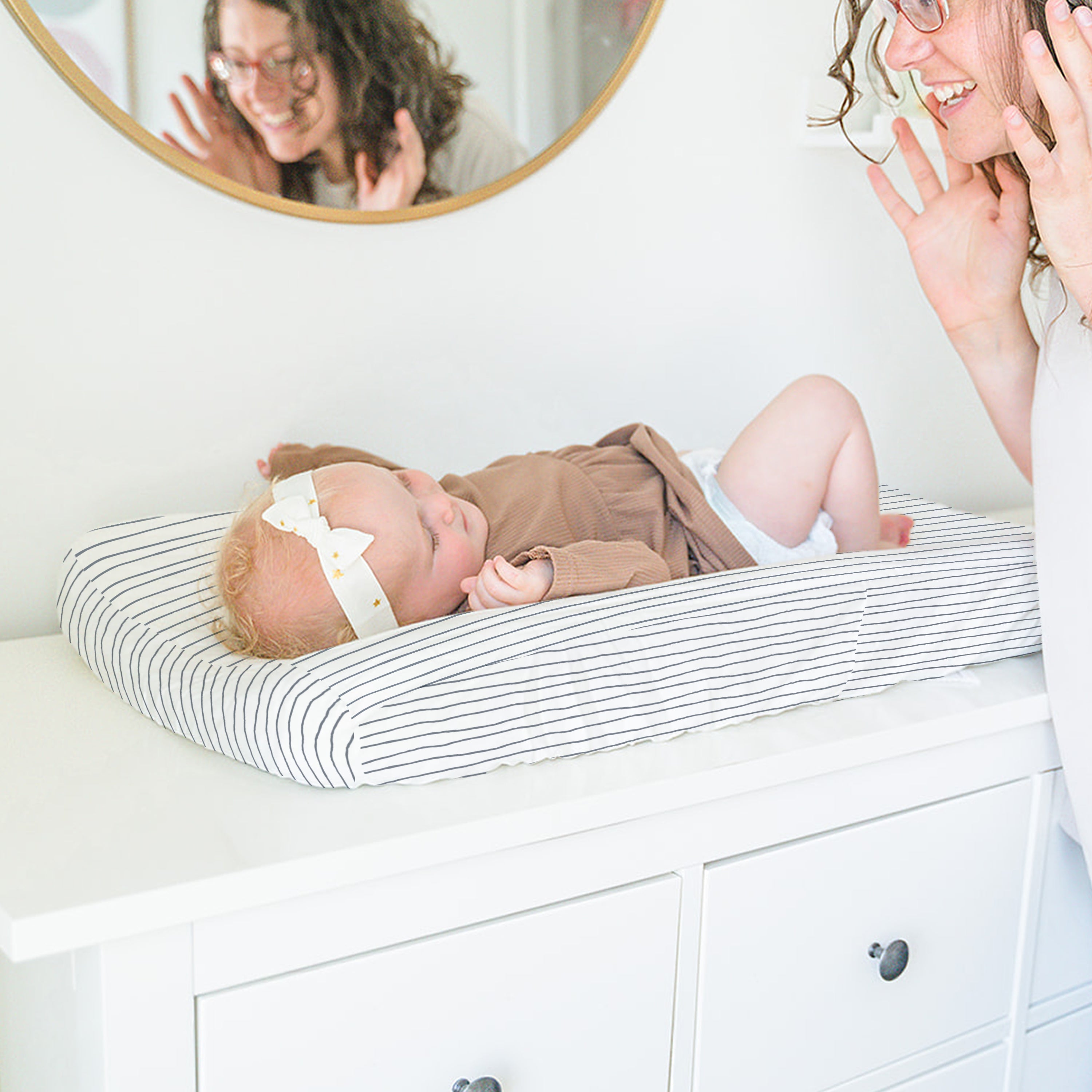 A joyful mother interacts playfully with her baby lying on a Makemake Organics Organic Cotton Changing Pad Cover - Navy Stripes, with their reflection visible in a circular mirror above them.