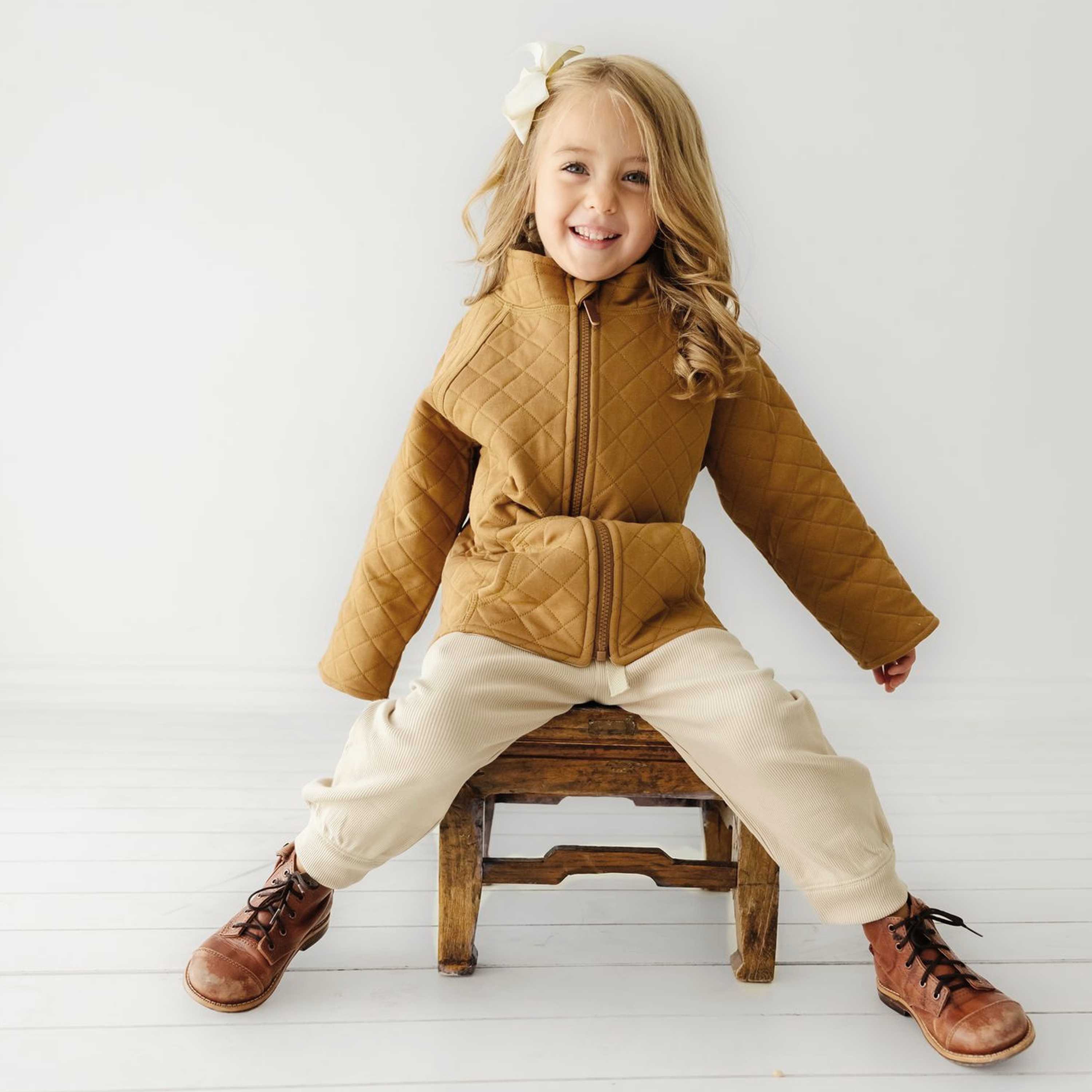 A joyful young girl wearing an Organic Kids Organic Merino Wool Zip Jacket in tan and cream pants, sitting on a rustic wooden stool against a white background, with a big smile and a white flower in her hair.