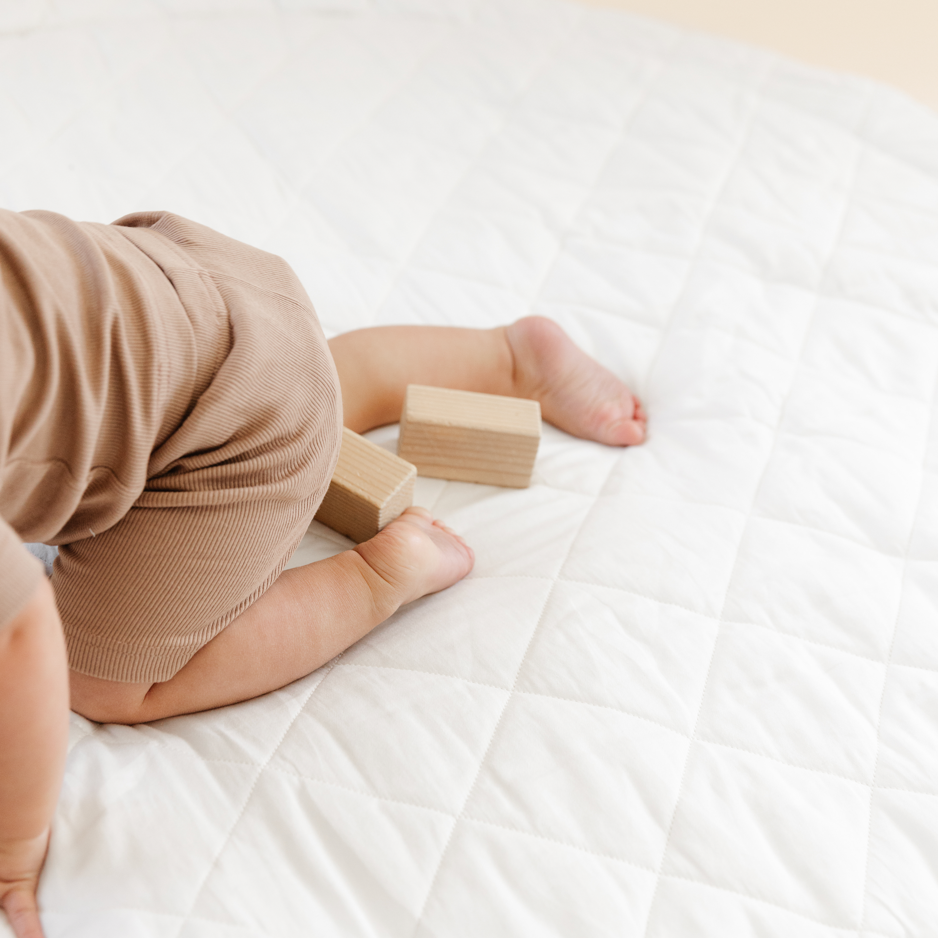 A baby in a brown onesie sits on an Organic Cotton Quilted Reversible Play Mat - Blossom  in Ivory, playing with two wooden blocks. the focus is on the baby's back and bare feet.