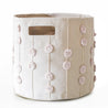 A cylindrical light beige fabric Makemake Organics Storage Basket Pompom Blush Oat adorned with decorative pink pom-poms strung vertically in rows, featuring a small cut-out handle on one side, isolated on a white background.