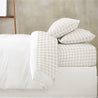 A neatly made bed with a white duvet and three pillows with beige and white gingham covers, set against a plain grey wall.