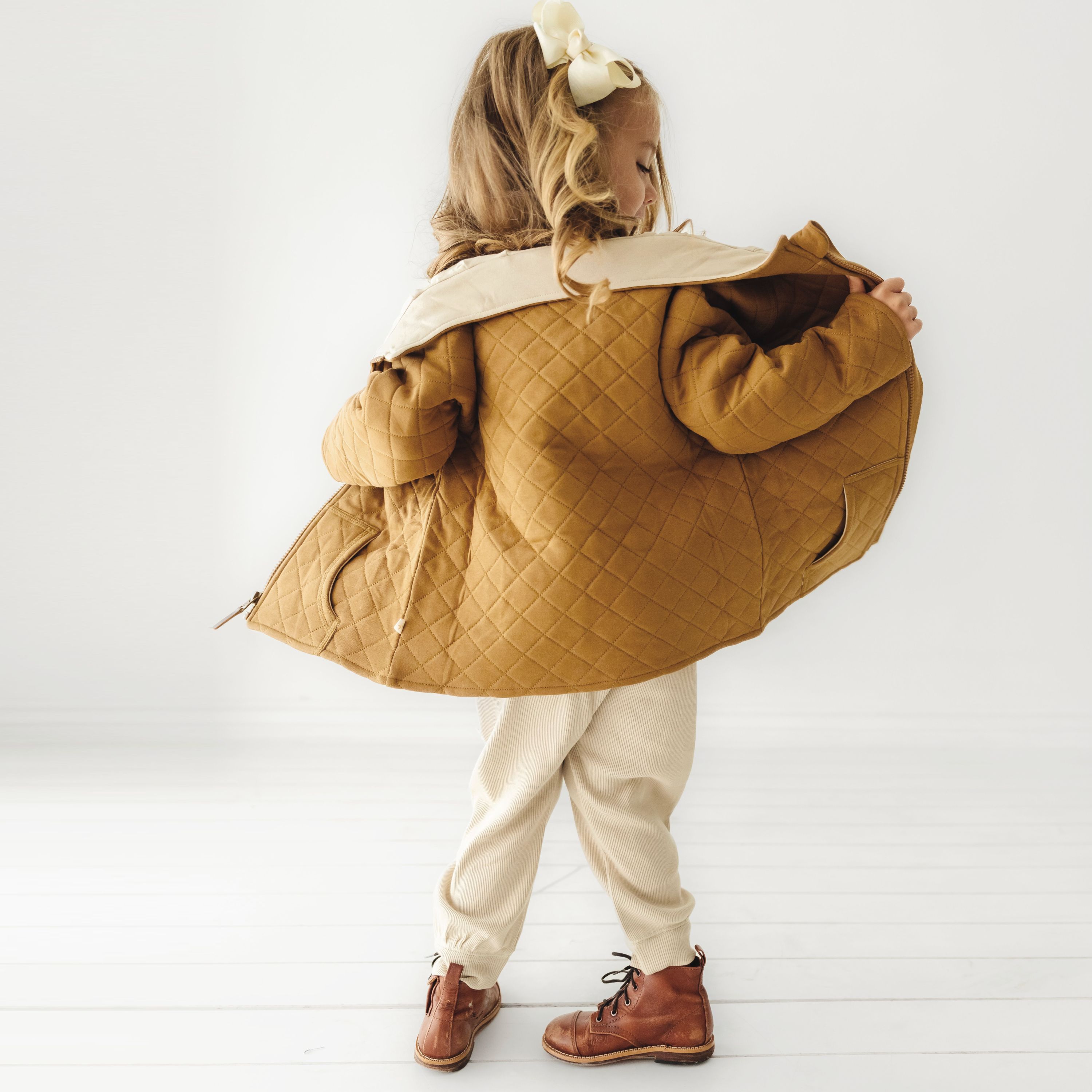A little girl with a ponytail, wearing a large Organic Kids tan merino wool zip jacket, beige pants, and brown boots, stands with her back to the camera in a white room.