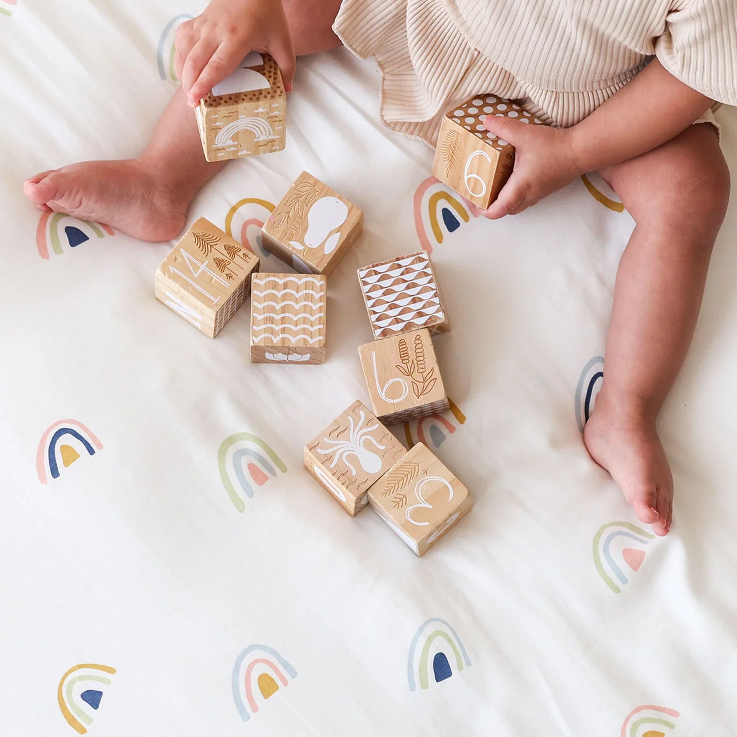 A toddler sits on a white Makemake Organics Mini Crib Fitted Sheet Set- Dotty & Rainbow with rainbow patterns, playing with wooden blocks featuring various carved designs. some blocks are scattered around the child.