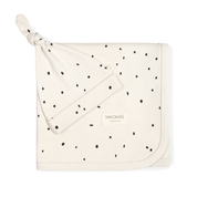 A cream-colored Makemake Organics swaddle blanket & hat neatly folded on a white background, decorated with a scattered black dot pattern, featuring a small visible logo.