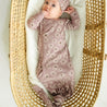 A toddler in a Organic Kimono Knotted Sleep Gown - Daisies by Makemake Organics lies in a woven basket with a soft liner, looking up with hands near face.